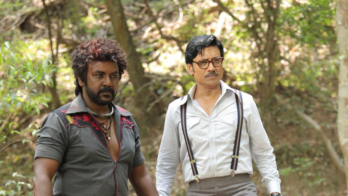 Raghava Lawrence, wearing a collared shirt unbuttoned at the top, and S.J. Suryah, wearing overalls over a white buttoned-up shirt, stand in a forest together in Jigarthanda DoubleX.